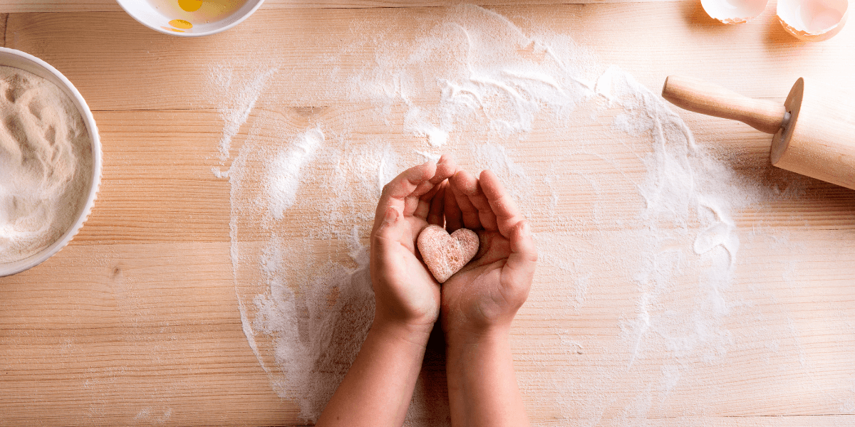 Hands on a floured table holding a heart shaped biscuit. rolling pin and ingredients surround them
