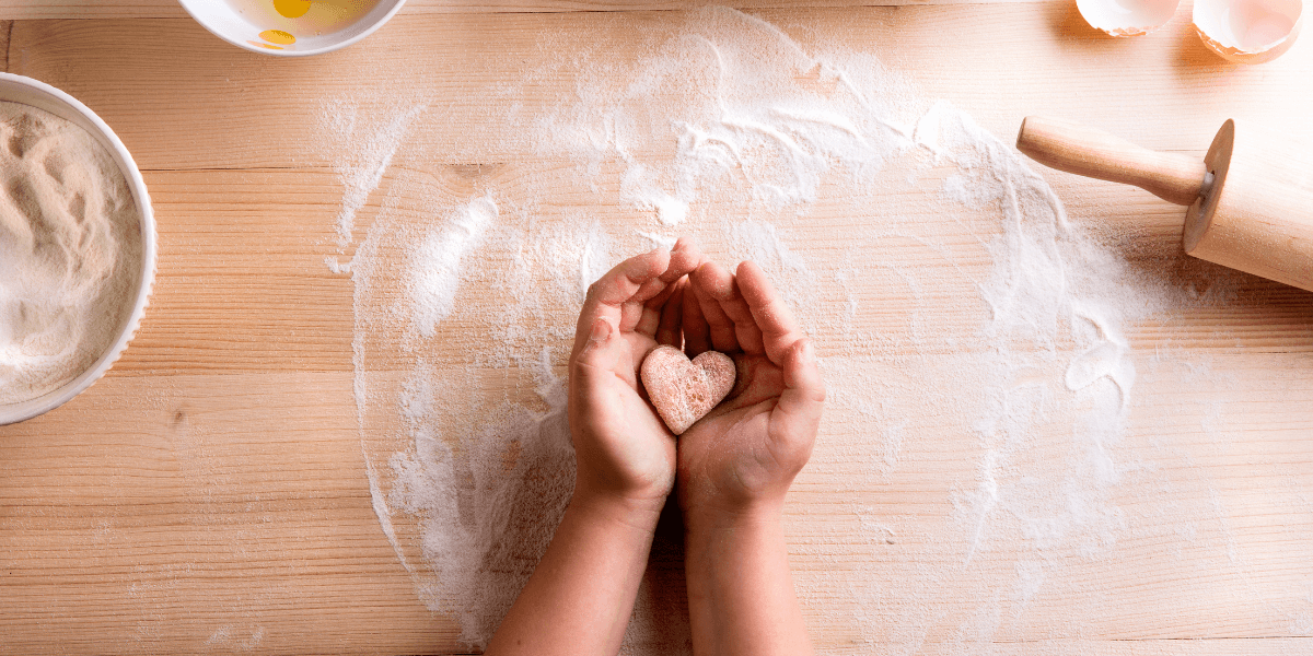 Hands on a floured table holding a heart shaped biscuit. rolling pin and ingredients surround them 